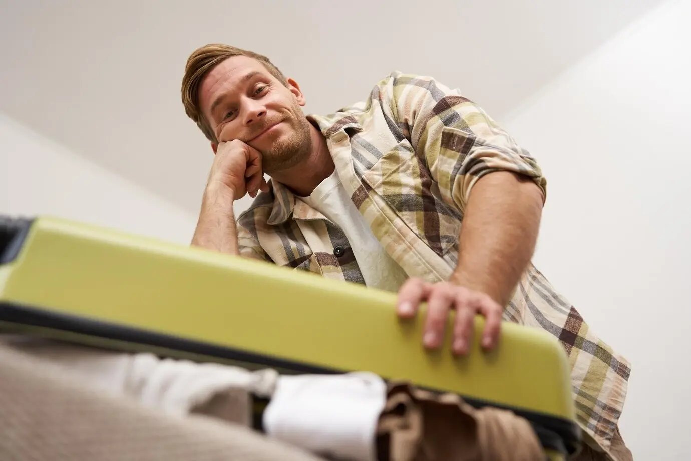 Image of a funny man trying to close a suitcase stuffed with a pile of clothes while packing luggage